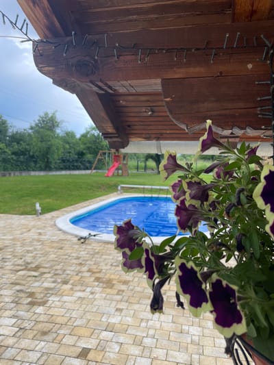 A photograph taken in Baevtsi, Bulgaria, shows a partially covered swimming pool, surrounded by light-colored stone pavers. The pool's cover is blue. A wooden structure with string lights is visible in the upper left corner. Purple and white flowers hang in the foreground, partially obscuring the view. In the background, a grassy lawn leads to a tree line, with a children's play structure including a red slide visible.