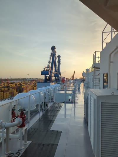 The image depicts a view from the deck of a large vessel, likely a cargo ship, at a port in Hambantota, Sri Lanka. The sun is low in the sky, casting a warm glow and creating long reflections on the wet deck. The foreground shows a white railing, several white cylindrical lifeboats covered with protective domes, and red fire hydrants with white piping. The deck itself is a light gray with darker rectangular sections.
In the midground, large blue port cranes dominate the scene, suggesting a busy container terminal. Beyond the cranes, stacks of yellow and red shipping containers are visible, stretching into the distance. Several lampposts are also present in the port area. The background consists of a pale, hazy sky with subtle clouds, indicating either sunrise or sunset. The overall impression is one of industrial activity and maritime operations in a coastal environment. There are no people visible in the image.