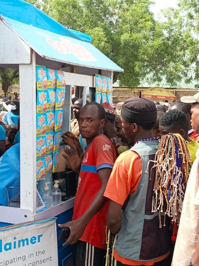 A medium shot captures a food vending stand in Kawaji, Nigeria, surrounded by a crowd. The vending stand, under a blue awning displaying "GOLDEN MORN," has packages of "GOLDEN MORN" cereal prominently displayed. A man in a red shirt with black-and-white striped pants gestures towards the stand with one hand and holds a phone in the other. Another man, wearing an orange shirt and a grey vest, has strands of colorful beads hanging on his back. The crowd behind them appears diverse, with people in various traditional and casual clothing. In the background, an umbrella with "MAGGI" branding is visible. The environment appears to be an outdoor public space.