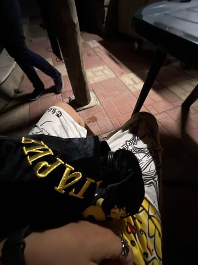 The scene is an outdoor, low-light environment in Debelt, Bulgaria, viewed from a first-person perspective looking downward. The ground consists of reddish-brown rectangular paving tiles.

In the immediate foreground, a hand is visible on the right, wearing a metallic ring with a complex design on the ring finger. Resting on the lap is a black velour-like garment with prominent gold embroidered text, partially reading "TRAP" or "TRAPSTAR." Beneath this, a white garment with black illustrative patterns is visible, along with a portion of a leg wearing yellow patterned shorts or pants with black outlines and scattered orange accents.

To the left, a bare leg wearing white shorts with dark textual or graphic designs is visible. A robust, vertical wooden beam or post stands upright on the tiled surface. Further into the midground, a dark, slender table leg supports a dark-colored, possibly plastic, tabletop, with only a segment visible on the upper right.

In the background, another individual is partially visible, walking away to the left. This person is wearing dark-colored trousers or jeans and dark sneakers with white soles. The illumination is dim, characteristic of night-time, resulting in prominent shadows cast across the tiled ground by the walking individual, the table, and the wooden post.
