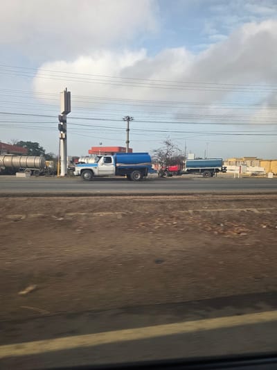 The image depicts a roadside scene in El Dátil, Venezuela. Several trucks of varying sizes and colors are parked along the shoulder of a paved road. Overhead power lines intersect the sky, which is partly cloudy. Low-rise buildings can be seen in the background. The foreground consists of a blurred roadside.