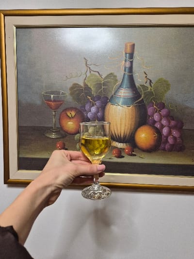 The image depicts an indoor scene within the Municipal Unit of Corinth, Greece. A light-colored wall serves as the background for a framed still-life painting. The painting features a gold-colored frame with an inner off-white mat and displays a composition including a dark blue, straw-wrapped bottle with a cork stopper, bearing a label that reads "Frizzante". Accompanying the bottle are bunches of purple grapes, red apples, orange-colored citrus fruits, and several red cherries, along with a stemmed glass containing a red liquid. In the foreground, a human hand, identifiable by light pink nail polish on the fingernails, extends from the lower-left. The hand grasps a clear, stemmed glass filled with a translucent yellow-gold liquid. This hand and glass are positioned in front of the lower-center of the painting, partially obscuring it. A dark fabric, consistent with a sleeve, is visible at the bottom-left edge of the frame. A faint shadow from the hand and glass is cast on the wall below them.