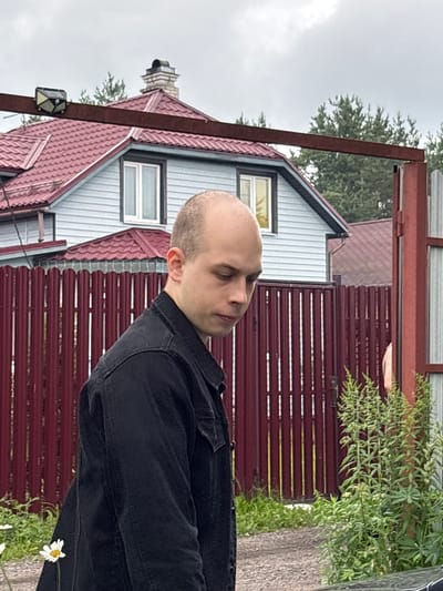 The image shows a young, bald man wearing a black denim jacket, standing in front of a red metal fence and a light blue house with a red roof. The house has a chimney. There are trees and some vegetation in the background, including daisies. The sky is overcast. The scene appears to be a residential area, likely in the city of Leningradskaya, Russia, given the architectural style and vegetation. The man is looking downwards and to his right, with his profile visible. The image captures an outdoor, possibly suburban, environment.