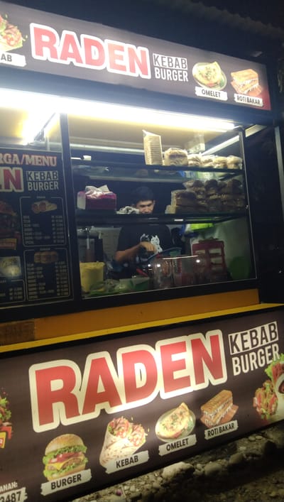 This image shows an outdoor food stall at night in Keude Trienggadeng, Indonesia. The stall's signage, illuminated by bright lights, prominently features the name "RADEN" in large red letters, with "KEBAB BURGER" written below. The sign also displays images of various food items like omelets and grilled bread.

Inside the stall, a young man wearing a dark t-shirt is visible behind a glass counter, appearing to be preparing food. Shelves behind him are stocked with bread, packaged items, and other ingredients. The scene suggests a busy food service operation, likely catering to late-night customers. The ground in front of the stall is rough and uneven, with some rocks visible, indicating a street-side or open-air location. The overall atmosphere is that of a bustling local eatery operating after dark. Visible text includes menu items and prices, along with the stall's name and product offerings.