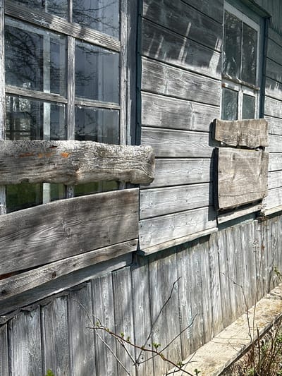 The image depicts a dilapidated structure in Студзёнкаўскі сельскі Савет, Belarus. The building appears to be constructed primarily of weathered wood. Multiple boarded-up windows are visible, with some window panes broken or missing. The wood exhibits signs of age and decay, including peeling paint and discoloration. The boards covering the windows are uneven and appear to be makeshift repairs. Some vegetation is visible on the right side of the frame, suggesting the structure is in a state of disuse and may be surrounded by nature. No people are visible in the frame.