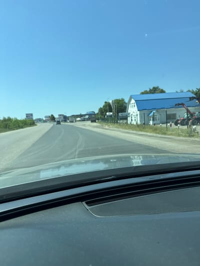 The media file depicts a street scene viewed from inside a vehicle. The image is oriented vertically, with the sky at the top and the ground at the bottom. In the background, several buildings with blue roofs are visible, along with some agricultural machinery. Trees and green foliage line the side of the road, suggesting a rural or semi-rural setting. The road itself is paved and appears to be a two-lane road. Several vehicles are present, including cars and what appear to be trucks or larger vehicles. The sky is clear and bright blue, indicating a sunny day. The overall impression is a snapshot of a typical day in a small town or agricultural area. There is no visible text in the image.