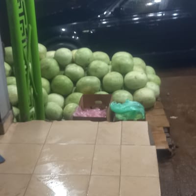 A stack of light green, melon-like fruits is stacked next to a tiled wall. To the left of the stack is a green object. A dark vehicle is visible in the background. The location is possibly a marketplace or retail setting in Barnawa, Nigeria.