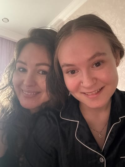 A close-up, rotated selfie features two women smiling cheerfully. They are positioned one above the other, with their faces filling most of the frame. Both women appear to be wearing black, satin-like pajama tops with white piping, suggesting a relaxed, indoor setting, likely in the evening or at night.

The woman positioned higher has light brown hair swept back from her face. She has warm brown eyes, a small stud piercing on her right nostril, and is smiling broadly, revealing her teeth. A delicate gold stud earring is visible in her right ear, and a thin gold-colored necklace with a small, circular or crescent-shaped pendant adorns her chest.

The woman positioned below has darker, wavy brown hair, and also has brown eyes. She is smiling widely, with her teeth visible, conveying a similar expression of happiness. Her face is partly shadowed by the hair of the woman above.

Both individuals exude happiness and warmth through their genuine smiles. The background is an indistinct, light-colored surface, possibly a wall or ceiling, with no specific identifying features visible beyond a faint circular object that may be a light fixture or decorative element. There is no visible text in the image.