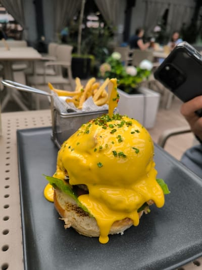 The image depicts a meal served on a light-colored, perforated outdoor table within a restaurant setting in Oława, Poland. In the foreground, a large, round food item, likely a burger or similar dish, is presented on a dark gray rectangular plate. This item is heavily covered in a thick, bright yellow sauce, possibly cheese or hollandaise, which thoroughly coats the top and drips down the sides, revealing green lettuce and a toasted bun beneath. A wooden skewer, also partially covered in the yellow sauce, is inserted vertically into the center, topped with chopped green chives and small red specks. To the left and slightly behind this dish, a metal basket holds a serving of golden-brown french fries contained within crumpled paper. On the right side of the frame, a partially visible hand holds a black smartphone with a multi-lens camera array, positioned as if capturing the food. The background is blurred, showing additional light-colored tables and chairs on what appears to be a patio or terrace, with indistinct figures seated at some tables. Potted plants with greenery and light-colored flowers, along with light-colored drapes, are visible in the soft-focus background, indicating an outdoor or semi-outdoor dining area.