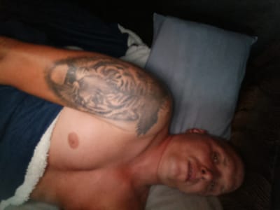 An individual is lying down, possibly on a couch or bed, with their head resting on a pillow. They have a tattoo of a tiger on their left arm. The person appears to be male and is wearing dark blue clothing with a white furry trim. The lighting is dim. The location in Rudamina, Lithuania, is unknown.