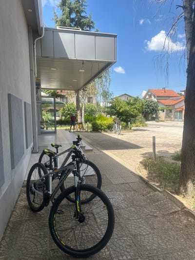 The image depicts an outdoor scene in the city of Ovoshtnik, Bulgaria, on a sunny day. In the foreground, two parked bicycles with silver frames and yellow accents are positioned on a tiled walkway adjacent to a light-colored building. The bicycles are facing towards the right of the frame. Further back, a person wearing a red shirt and shorts is visible near an open doorway, interacting with a dog. A light blue bicycle is parked in the background to the left, near some greenery and a bike rack. The environment includes trees, houses with red-tiled roofs, and a clear blue sky with scattered clouds. The ground is a mixture of pavement, dirt, and sparse vegetation.