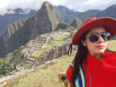 A woman in a red hat and colorful poncho poses in front of the ancient Inca citadel of Machu Picchu. She wears aviator sunglasses, reflecting the surrounding landscape. The ruins are spread across the mountainside, with prominent stone structures and grassy terraces. The background features a steep, cloud-draped mountain and distant peaks, creating a scenic panorama. A small wooden sign with an arrow is visible at the bottom.