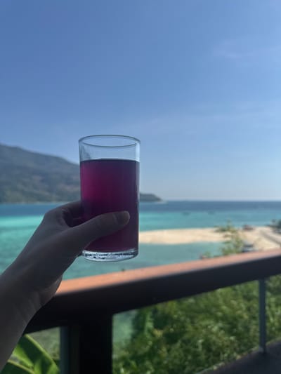 An elevated view captures a human hand holding a clear, cylindrical glass filled with a magenta-purple liquid, showing condensation droplets. The hand grips the lower portion of the glass, presenting it towards the viewer. In the immediate foreground, a dark wooden railing and green foliage are visible. The background features a panoramic tropical seascape of Ban Ko Li Pe, Thailand, under a clear blue sky. A light-colored sandy beach or sandbar separates vibrant turquoise shallow water from deeper blue ocean, where several small boats are scattered. A large, verdant landmass or island rises from the sea on the left horizon. The scene is brightly lit by natural sunlight.