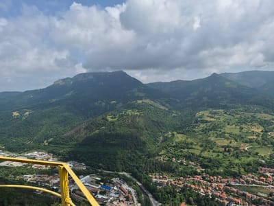 This is a high-angle, wide shot of a mountainous landscape with a town nestled in a valley. In the foreground, a bright yellow railing is visible, suggesting the viewpoint is from an elevated structure or observation deck. The railing is metallic and has a chain connecting two sections.
The middle ground features the town of Teteven, Bulgaria, with its characteristic red-tiled roofs. The town appears to be spread out along a winding road and a river or stream. Industrial buildings with flat roofs, some with solar panels, are clustered in the lower left portion of the image, adjacent to the residential areas.
The background is dominated by rolling green mountains covered in dense forest. The peaks are partially obscured by dramatic, overcast clouds. The sky is a mix of blue and grey, with puffy white clouds scattered across the expanse. The overall impression is one of a rural and scenic location. There are no people visible in the image.