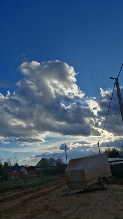 The image shows a cloudy sky above a rural scene. On the right side, a trailer sits on a dirt surface. In the background, there are houses and vegetation. Overhead power lines are visible, and the location is Otchor, Russia.