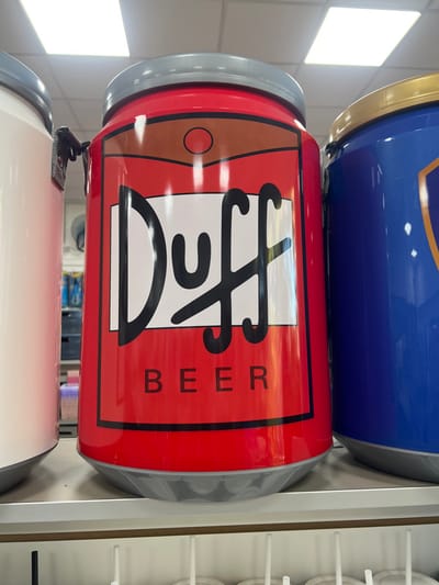 The image displays three large, can-shaped insulated containers or novelty items positioned on a white retail shelf within an indoor commercial establishment. The central object is a red cylindrical container, approximately 60-70 cm in height, featuring a label with "Duff" in black stylized script and "BEER" in black capital letters below it on a white rectangular background. This container has a gray top lid and a gray base. To its left, a partial view of a white cylindrical container with a gray lid is visible, exhibiting a black strap or handle attachment near its top. To the right, a partial view of a blue cylindrical container with a gold-colored lid is visible. The background reveals a retail environment with overhead fluorescent lighting panels on a white tiled ceiling and blurred displays of various merchandise. No individuals are present. No specific visual cues within the image permit the precise identification of its location within the city of Santa Rosa, Argentina.