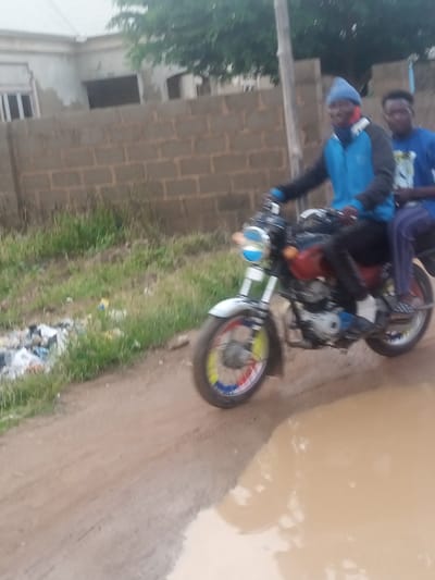 The image depicts two young men riding a motorcycle on a dirt road in what appears to be Russo, Nigeria. The driver, in the foreground, wears a blue beanie and a blue and black jacket. He is looking towards the camera with a slight smile. The passenger, behind him, wears a blue t-shirt. They are both wearing dark pants.

The motorcycle is red and silver with colorful accents on the wheels, featuring red, white, blue, and yellow. The scene appears to be outdoors, with a brick wall in the background. To the left of the road, there is overgrown grass and some scattered trash. The road itself is dirt and muddy, with a large puddle in the foreground, suggesting recent rain. The sky is overcast, indicating a cloudy or potentially rainy day. The overall lighting suggests it is daytime, though the weather conditions are making it appear somewhat dim. The context of Russo, Nigeria, places the scene in a specific geographical and cultural setting. No visible text is present in the image.