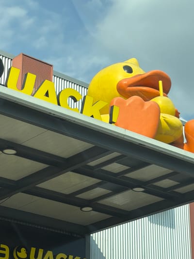 The image shows a large, stylized yellow duck figure with an open orange beak holding a yellow popsicle stick, mounted on a building with the name "QUACK". The building has a modern architectural design with a dark-colored crossbeam pattern. The sky with clouds is visible behind the duck. The photograph was taken from a low angle. This is the famous “Pollos Quack” restaurant in San Pedro Cholula, Mexico.