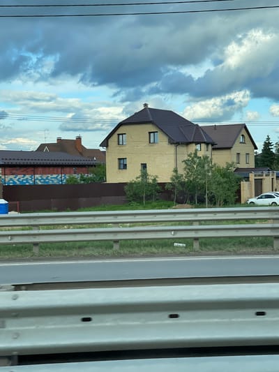 The image shows a residential area in Ivlevo, Russia. Two detached houses are visible: one is two stories with a dark brown roof and light yellow brick walls, and the other is partially obscured but appears to be similar in style. A white car is parked near the houses. A fence, partly brown and partly patterned with blue and red, divides the properties. A road barrier with metal bars runs along the right side of the frame. Overhead power lines are visible against a cloudy sky.