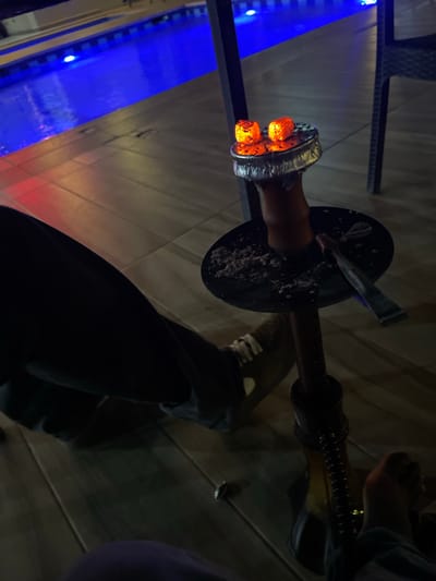 A partially obscured hookah is the central subject, with lit coals in the bowl. The hookah is positioned on a wooden surface, presumably a table, with a dark metallic tray underneath. A leg with a grey and white shoe is seen partially in the foreground. The background features a blue decorative light feature, possibly a water wall. The location is reported to be Rubuchi, Nigeria, although no specific identifying landmarks are visible in the image to verify the exact locale. The interaction appears to be someone preparing or engaging in hookah smoking.