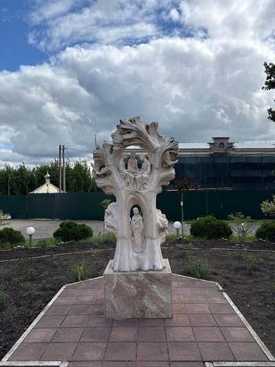 The image depicts a white stone sculpture of a tree with figures carved within its trunk and branches, set on a marble pedestal. The sculpture is positioned in a garden bed with dark soil, surrounded by a paved walkway. In the background, a green metal fence separates the garden from a street with trees and power lines. Further back, a domed building with a golden roof, possibly a church, is visible. Beyond that, a large building with scaffolding and dark netting indicates ongoing construction or renovation. The sky is partially cloudy with a mix of blue sky and white clouds. The overall scene suggests an outdoor public space or religious grounds.