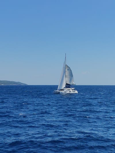 A white catamaran sailboat is sailing on a deep blue ocean under a clear, pale blue sky. The catamaran has two hulls and is equipped with a tall mast and white sails that are partially unfurled. The boat is positioned in the right half of the frame, moving slightly to the left. In the distance, on the left side of the image, a green, tree-covered island is visible along the horizon. The ocean surface is choppy with visible waves. The scene appears to be a bright, sunny day.
