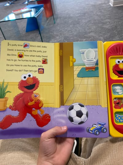 The image shows an open children's book on a patterned floor. The left page has text and illustrations related to potty training, featuring characters Elmo and baby David. Elmo is depicted holding a baby, running. The right page is a part of an interactive toy, with buttons and images of Elmo. In the foreground, a person's hand is visible, holding the book open. The background shows a blurred interior, possibly a living room with glass furniture. The scene appears to be indoors, with no indication of its specific location in Chesapeake, United States of America.