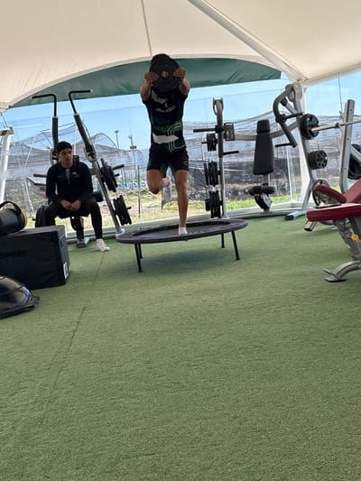 Inside a gym, a man balances on a trampoline, holding a weight plate in front of his face. He wears a t-shirt and shorts. Another man in a black jacket sits on a weight machine nearby. Exercise equipment surrounds them, including weight racks and a bench press. The gym has an artificial turf floor and a transparent, tent-like roof.