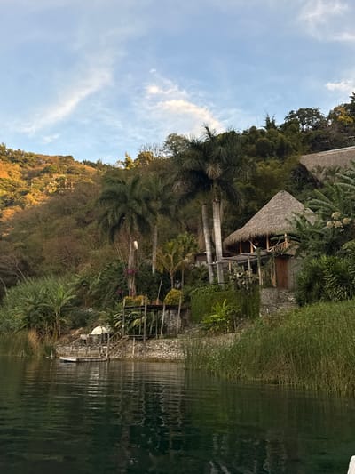 The image captures a tranquil lakeside scene in San Pablo La Laguna, Guatemala. The foreground consists of dark green, calm lake water with slight surface ripples reflecting the sky. The immediate shoreline is defined by a stone retaining wall. On the left, a small wooden floating dock extends into the water, with wooden steps ascending to a paved area where a small white dome-shaped structure and a wooden trellis-like stand with potted plants are visible. To the right, tall green reeds grow densely along the water's edge.

Above the shoreline, a steep hillside rises, covered in lush tropical vegetation including palm trees, broad-leafed plants, and dense shrubs. Prominently situated on the hillside is a two-story building featuring a steeply pitched thatched roof. Its base appears to be stone, while the upper level includes an open-air balcony or patio with wooden railings, from which a hammock is suspended. A wooden door is visible on its right side. To the right and slightly behind this structure, a second building with a similar thatched roof is partially visible, nestled further up the slope amidst the trees. Scattered throughout the hillside are various palm trees and other dense foliage.

The background reveals a larger, verdant mountain or hill range, with some areas illuminated by golden sunlight, suggesting either early morning or late afternoon. The sky above is clear blue with sparse, wispy white clouds. No human figures or animals are present in the scene.