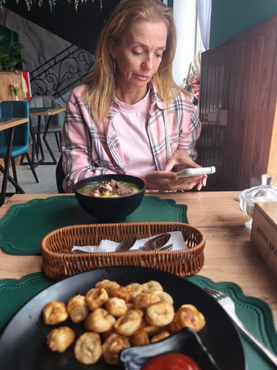 A woman with light brown hair, wearing a pink t-shirt and a pink and white plaid shirt, sits at a table in a restaurant. She is holding a white smartphone and looking at it. In front of her are two plates of food. The first plate contains soup in a black bowl. The second plate is a black plate containing fried dumplings and a small black dish with red sauce. A brown wicker basket with silverware is between the two plates. The table is wooden with green placemats. In the background are chairs and a staircase. The location is Semenovka, Kyrgyzstan.