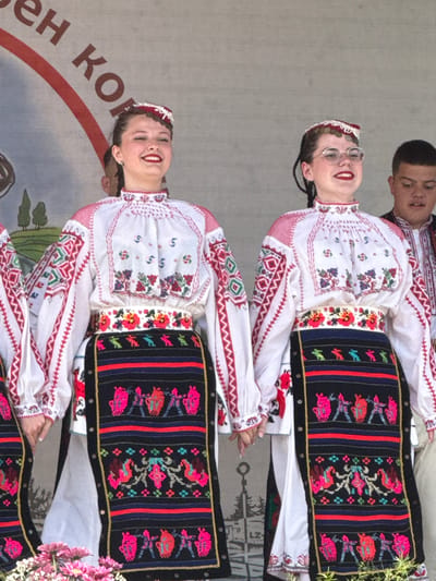 This is a photograph taken in the city of Pordim, Bulgaria, possibly during a cultural event. The image focuses on two young women dressed in traditional Bulgarian folk costumes. Both women are wearing white blouses with intricate red and colorful embroidery on the collars, cuffs, and chests. They also wear decorative sashes around their waists, adorned with floral patterns and geometric designs. Their skirts are black and heavily embroidered with vibrant patterns, featuring figures and floral motifs in pink, red, blue, and green. Both women have their hair styled in braids and are wearing traditional headwear. The woman on the left has a red and white embroidered cap, while the woman on the right wears glasses and a similar embroidered cap. Behind them, another young man in traditional attire is partially visible. The background appears to be a stage or backdrop with lettering that is partially obscured, but suggests it might be related to an event or organization. The overall scene depicts a cultural celebration with a focus on traditional Bulgarian attire.