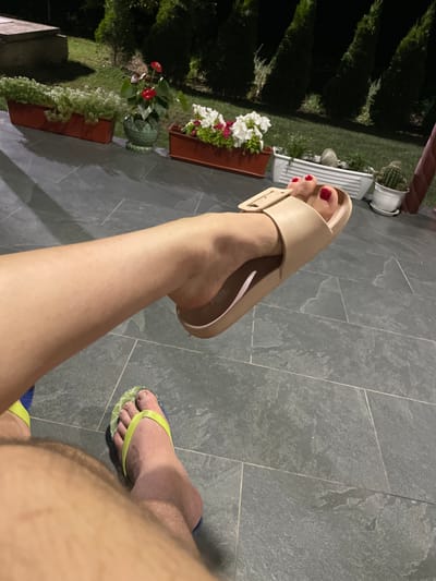 In Slobozia-Dușca, Moldova, a low-angle, outdoor, nighttime photograph captures two individuals' feet resting on a grey tiled patio. One person's left leg is prominently featured, bent at the knee, with their foot adorned in a tan slide sandal and red nail polish. To the left of this, the other individual's right foot is visible, wearing a green and blue flip-flop. The background reveals a manicured lawn, several potted plants including flowers and a cactus, and a line of evergreen trees, suggesting a residential or garden setting.