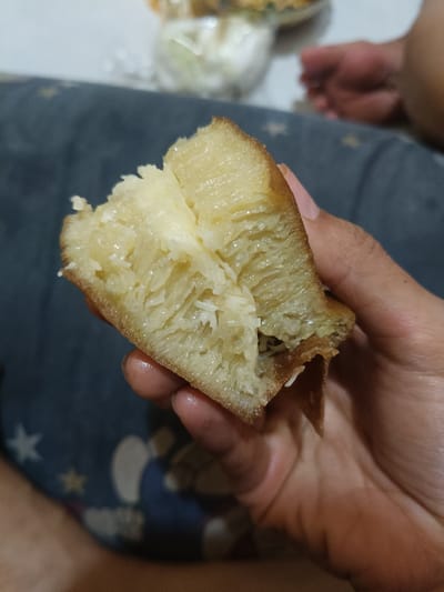 A human hand with medium-brown skin holds a freshly torn piece of Martabak Manis (sweet pancake) in an indoor setting located in Cileungsi, Indonesia. The pancake displays a golden-brown, slightly toasted crust on its exterior edges and a pale yellow, porous interior with a distinct honeycomb texture. Shredded white coconut is visible as a prominent filling within the open, torn section of the pancake, which also appears moist and slightly glossy, suggesting butter or oil. The hand grips the food item with the thumb, index, and middle fingers. The immediate background consists of a dark blue fabric surface adorned with white and yellow star patterns, along with blurry light-colored shapes. Further in the out-of-focus background, a white, indistinct container or object is present on the left, while a blurred light-brown human foot or hand is partially visible on the right. The overall environment is an informally lit interior space.