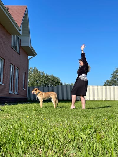 A woman in a black dress and a black hat is in mid-air, appearing to fly or jump, with her arms outstretched. She is on a grassy lawn, with a brick house to her left and a fence in the background. A dog, possibly a golden retriever mix, is standing on the grass in front of her, looking towards the right. The sky is a clear blue, indicating it is daytime with sunny weather. The scene appears to be a residential area, given the presence of the house and fence. The woman's pose suggests a moment of playfulness or a staged photograph. There is no visible text in the image.