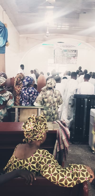 The image shows the interior of St. John's Anglican Church in Kaduna South, Nigeria, during a service. A congregation of people, many wearing traditional attire with vibrant patterns and headwraps, are seated on pews. In the foreground, a woman with a patterned headwrap and dress is seen from behind, resting her arm on the pew. The pews are made of dark wood. Behind the congregants, a banner hangs from the wall, advertising the "Harvest of Supernatural Increase." The room has a tiled ceiling with ceiling fans and fluorescent lighting. A man in a white garment stands facing the congregation in the background. The overall atmosphere appears to be one of communal worship and gathering.