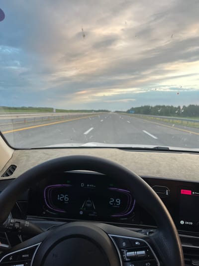 The image shows a view from inside a car driving on a highway. The dashboard displays a speed of 126 km/h and an engine RPM of 2.9 x1000rpm. The exterior view shows a multi-lane highway stretching into the distance under a cloudy sky with hints of sunset. The road is flanked by a guardrail and trees. The windshield has several smudges and specks, some of which appear to be red. The time displayed on the dashboard is 04:09. The location is identified as Altyn'dy Rural District, Kazakhstan.
