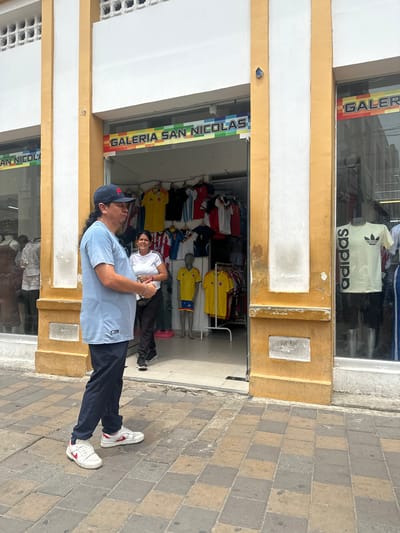 A man wearing a light blue t-shirt, navy pants, a baseball cap, and white sneakers with red accents stands on a paved sidewalk in front of a store. The store, labeled "GALERIA SAN NICOLAS," sells sportswear, with several jerseys visible on display inside. To the right of the entrance, a woman in a white t-shirt and black pants stands near a display of merchandise. The store facade is yellow and white, with large glass windows showcasing clothing. The pavement is composed of rectangular grey and tan tiles. In the background, other store fronts and buildings are visible. The scene is set in Perímetro Urbano Barranquilla, Colombia.
