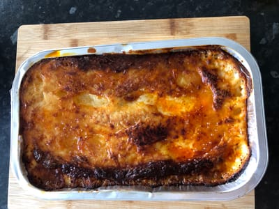 A close-up, top-down shot shows a rectangular foil tray filled with baked food, likely a gratin or casserole, resting on a wooden cutting board. The food is topped with a golden-brown, slightly charred cheese crust, with visible layers of mashed potato or pasta beneath. The tray's foil edges are slightly crinkled, and there are some dark, burnt sections on the perimeter of the food. The background is a dark, speckled surface, possibly a countertop. The image was taken at an undisclosed location in Hinckley and Bosworth, United Kingdom.