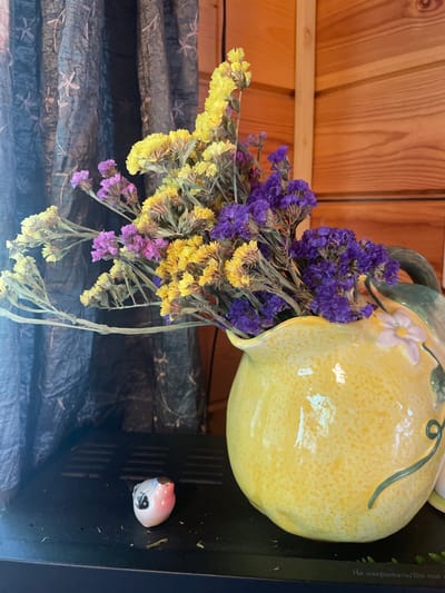 The image displays a close-up of a still life arrangement, likely in a domestic setting in Турово, Russia. In the foreground, a small, pink and white ceramic figurine resembling a bird or creature with black eyes is positioned on a dark, textured surface. To the right, a bright yellow, lemon-shaped ceramic pitcher holds a bouquet of dried flowers. The flowers are a mix of yellow and purple, with delicate, small blooms and dry, wispy stems. The pitcher has a light green decorative vine and a small pink and white flower embellishment. Behind the pitcher and flowers, a dark, textured fabric, possibly a curtain, hangs to the left, while a light-colored wooden wall paneled with horizontal planks forms the background on the right. The lighting appears natural, casting soft shadows. The scene suggests a decorative display indoors, possibly in a room with wooden walls.
