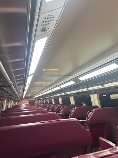 The image depicts the interior of a passenger train car. While the prompt specifies the location as Weston, United States, no visual cues within the image confirm this geographic detail. The perspective is down a central aisle, extending from a low vantage point towards the rear of the car. The ceiling consists of light gray panels, featuring rows of rectangular LED light fixtures providing bright illumination. A circular speaker grille and a small cylindrical sensor are visible on the ceiling toward the upper left. Multiple "EMERGENCY EXIT →" signs are affixed to the ceiling panels, indicating egress routes.
The train car contains numerous rows of seating upholstered in a deep magenta or plum color. These seats are arranged in opposing pairs along both sides of the aisle. One individual is visible on the right side of the car, several rows back from the foreground. This person is seated, facing towards the left (away from the window), with only their head visible in profile. The windows along the sides of the car reveal a dark exterior, indicating either nighttime or transit through a tunnel. Metallic luggage racks are situated above the windows. The overall scene portrays a sparsely occupied, well-lit, and orderly train compartment.