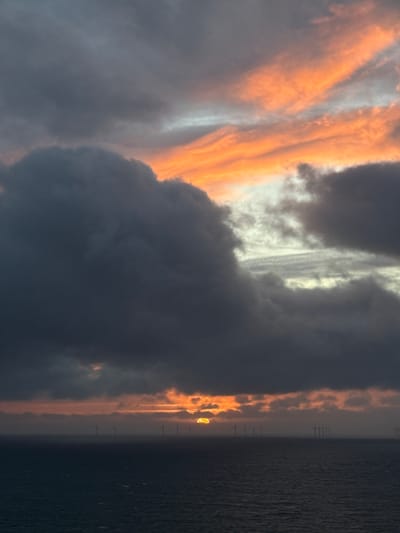 The image captures a dramatic sunset or sunrise over the sea, featuring an offshore wind farm on the horizon. The sky, which occupies the majority of the frame, is filled with an impressive array of clouds. On the left side, the clouds are intensely illuminated by the low-lying sun, showcasing vivid streaks of orange, yellow, and reddish hues that blend into soft whites and light grays, suggesting complex atmospheric layering. Towards the center and right, the clouds become denser and darker, appearing in deep grays and bluish-blacks, indicating thick storm clouds or areas where the light is entirely obscured.
The horizon line spans across the lower portion of the image, separating the dynamic sky from the water. The sea, visible on the right, is a dark, muted blue-grey, with a gently textured surface. Along the horizon, numerous tall structures, identifiable as wind turbines from an offshore farm, stand silhouetted against a narrow band of golden light. The sun's intense glow, possibly a partial view or a bright reflection, can be seen just above the horizon, peeking through a gap in the clouds and casting a brilliant yellow-orange light that highlights the cloud edges and reflects faintly on the water.
The scene conveys a powerful and awe-inspiring natural moment, characterized by the dramatic interplay of light and shadow across the vast sky and sea. The presence of an offshore wind farm aligns with the provided location context of Fécamp, France. No people or visible text are present in the image.