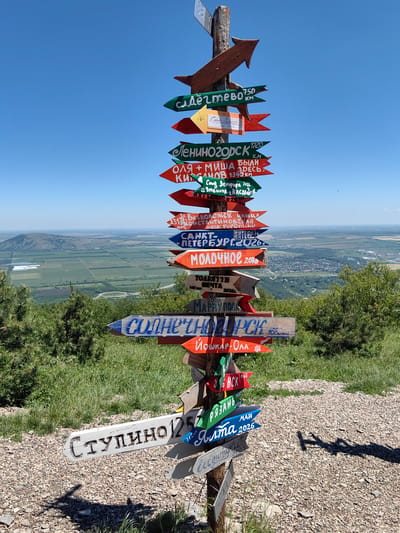 In Goryachevodsky, Russia, a wooden signpost stands on a gravelly, grassy hillside under a clear blue sky. The signpost is laden with multiple directional signs, each pointing in various directions and distances. The signs are hand-painted with different colored text, many indicating destinations and some with dates. Notable destinations include "Сляднево 750 км," "Лениногорск 1720," "Оля + Миша были здесь," "Санкт-Петербург 2026," "Молочное 2016," "Солнечный 2026," "Йошкар-Ола," "Рязань," "Ялта 2026," and "Ступино 1257." In the background, a vast, green valley stretches out, dotted with fields and a distant town, with rolling hills and a large mountain in the far distance. Trees and shrubs are visible on the hillside surrounding the signpost. No people are present in the image, and the scene appears to be a public outdoor landmark.