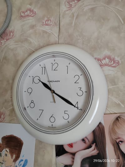 A white analog clock with a white frame is mounted on a wall with floral wallpaper in shades of beige and pink. The clock face is white with black Arabic numerals. The hour hand points to the number 10, and the minute hand points to the number 11. The second hand is thin and black, and it points to the number 3. The brand "TROYKA" and "QUARTZ" are visible on the clock face. Below the clock, two magazine covers are partially visible. The cover on the left features a drawing of two people. The cover on the right displays the faces of two women. A timestamp in the lower right corner reads "29/06/2026 10:22". The environment appears to be an indoor setting, likely a residential room. The presence of a cable near the top left of the image suggests it could be connected to the clock or another device.