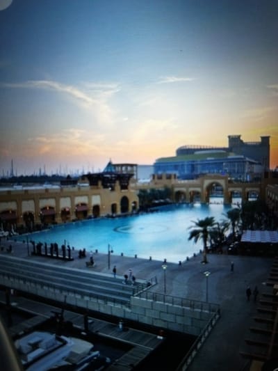 The image depicts an outdoor scene at dusk, with a large swimming pool in the foreground surrounded by buildings and walkways. The sky is a gradient of blue and orange, indicating sunset. In the distance, there is a modern building with a green roof. Several palm trees are scattered around the pool area, and there are people walking and standing on the walkways. The scene suggests a resort or entertainment complex. The provided location of Golbembal, Nigeria, is not supported by any visual evidence in the image.