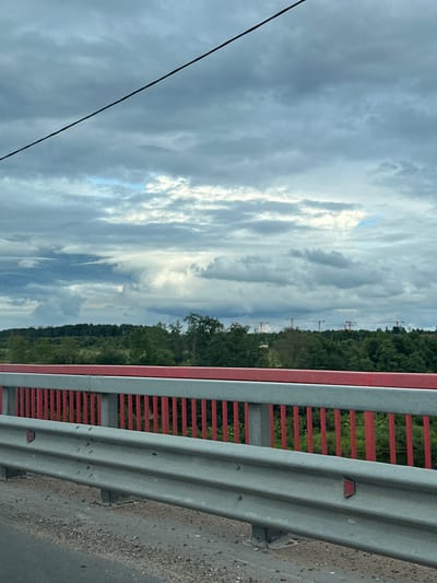 The image shows a view from a bridge in Kingisepp, Russia, looking towards a treeline under a cloudy sky. The bridge railing consists of red vertical posts and a grey horizontal bar, with a metal guardrail in the foreground. In the distance, beyond the trees, several construction cranes are visible. The sky is filled with dark, heavy clouds, with some lighter patches indicating potential breaks. The overall scene suggests an overcast day in a semi-urban or suburban area with ongoing construction.