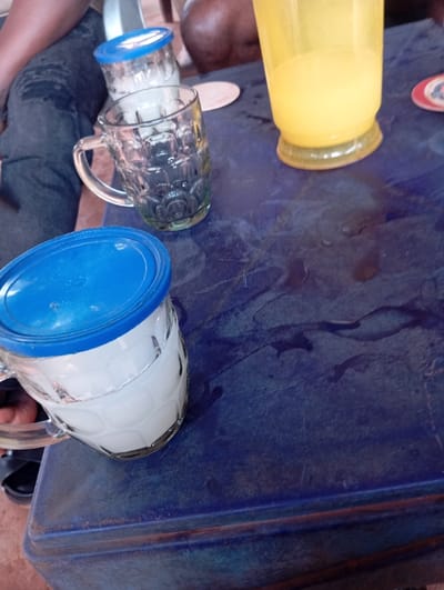 The image displays a close-up, high-angle view of a dark blue plastic table located in Okpatu-Ukehe, Nigeria. On the table, four beverage containers are present. In the lower left, a clear glass mug with a blue plastic lid contains a white opaque liquid. Centered on the table, a clear, empty glass mug with a patterned surface is visible. Behind it, a second clear glass mug with a blue plastic lid, also containing a white opaque liquid, is partially obscured. To the upper right, a yellow plastic cup holds a yellow opaque liquid. The table surface exhibits dark liquid stains and moisture. Two circular coasters are discernible: one light-colored and one with a red and white pattern. In the background, the lower body and legs of at least two individuals are visible; one wears dark blue jeans and black sandals, and another shows a dark-skinned thigh. A reddish-brown ground surface is visible beneath the table and individuals.