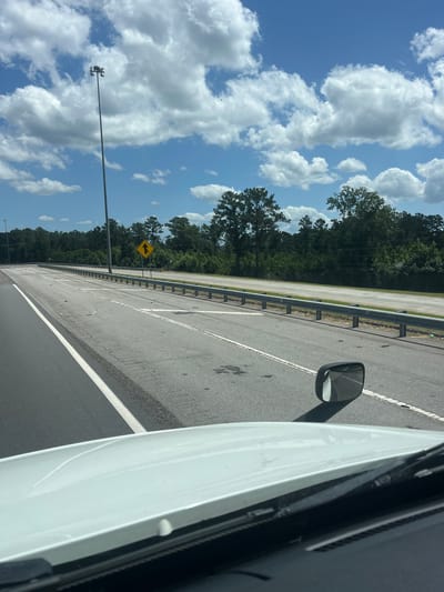 The image depicts a highway scene in Riceboro, United States, under a partly cloudy sky. The perspective is from inside a white vehicle, showing the hood and windshield wipers. The highway itself has multiple lanes, marked with white lines. A metal guardrail runs along the side of the road, separating it from a wooded area in the background. A tall light pole with multiple lamps is visible on the left side of the highway. A yellow diamond-shaped road sign with an arrow pointing upwards is positioned near the guardrail. Scattered debris can be seen on the road surface. A side mirror of the vehicle is also visible, reflecting the highway and the sky.