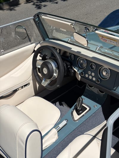 A high-angle, close-up shot captures the interior of a vintage-style convertible car, possibly a Morgan, parked outdoors. The vehicle's interior features light-colored leather seats with dark blue piping, a black leather-wrapped steering wheel with a silver emblem in the center, and a dashboard with a classic design. The dashboard is equipped with multiple analog gauges, buttons, and a prominent gear shifter. Sunlight streams in, casting shadows across the interior and the surrounding asphalt, suggesting a sunny day. The background is blurred, but hints of trees and a street indicate an outdoor urban or suburban setting, consistent with Village of Sag Harbor, United States of America. No people are visible in the image.