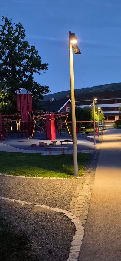 The image captures a playground at dusk in Lillehammer, Norway. A tall wooden lamppost with two lights stands prominently in the foreground, illuminating a path paved with asphalt and edged with cobblestones. To the left, a red playground structure with slides and climbing frames is partially visible, set against a backdrop of trees and a dark blue sky. In the distance, a red house with white trim and a dark roof is nestled amongst trees, with a sloping hill in the background. The playground appears deserted, with no people present. The lighting suggests it is either early evening or early morning, with artificial lights from the lampposts casting a warm glow on the surroundings.