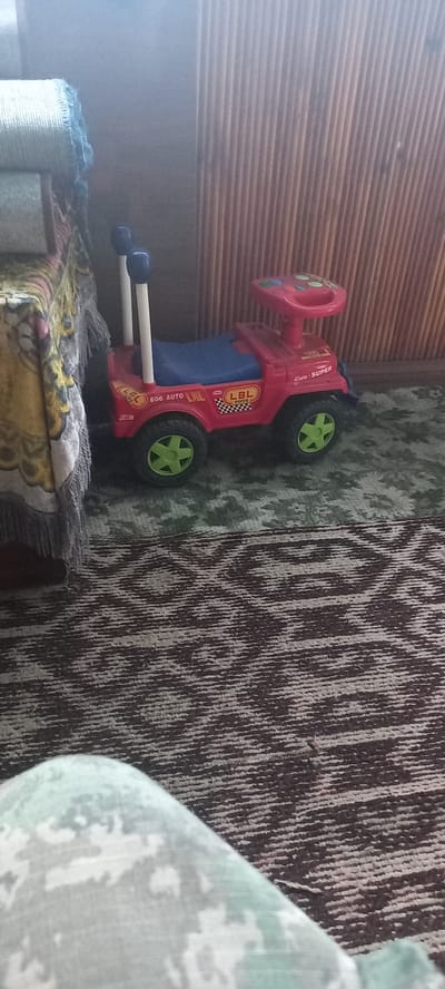 A red and blue ride-on toy car is positioned on a carpeted floor next to a draped couch. The car has green wheels and a steering wheel on top. A person's leg, wearing camouflage-patterned clothing, is visible in the lower foreground. The background consists of a wall with vertical wooden slats. This scene appears to be indoors, likely in a residential setting in the village of Tokushi, Kazakhstan.